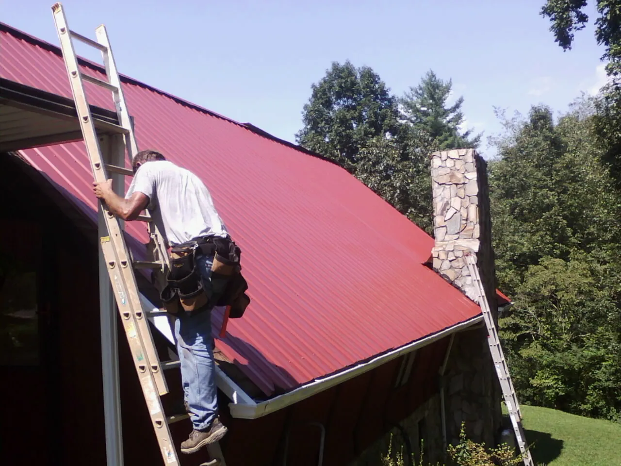 Roofer on a ladder beside a red metal roof and stone chimney — Soffit & Fascia Repair by Roofing Brothers in Oriskany Falls
